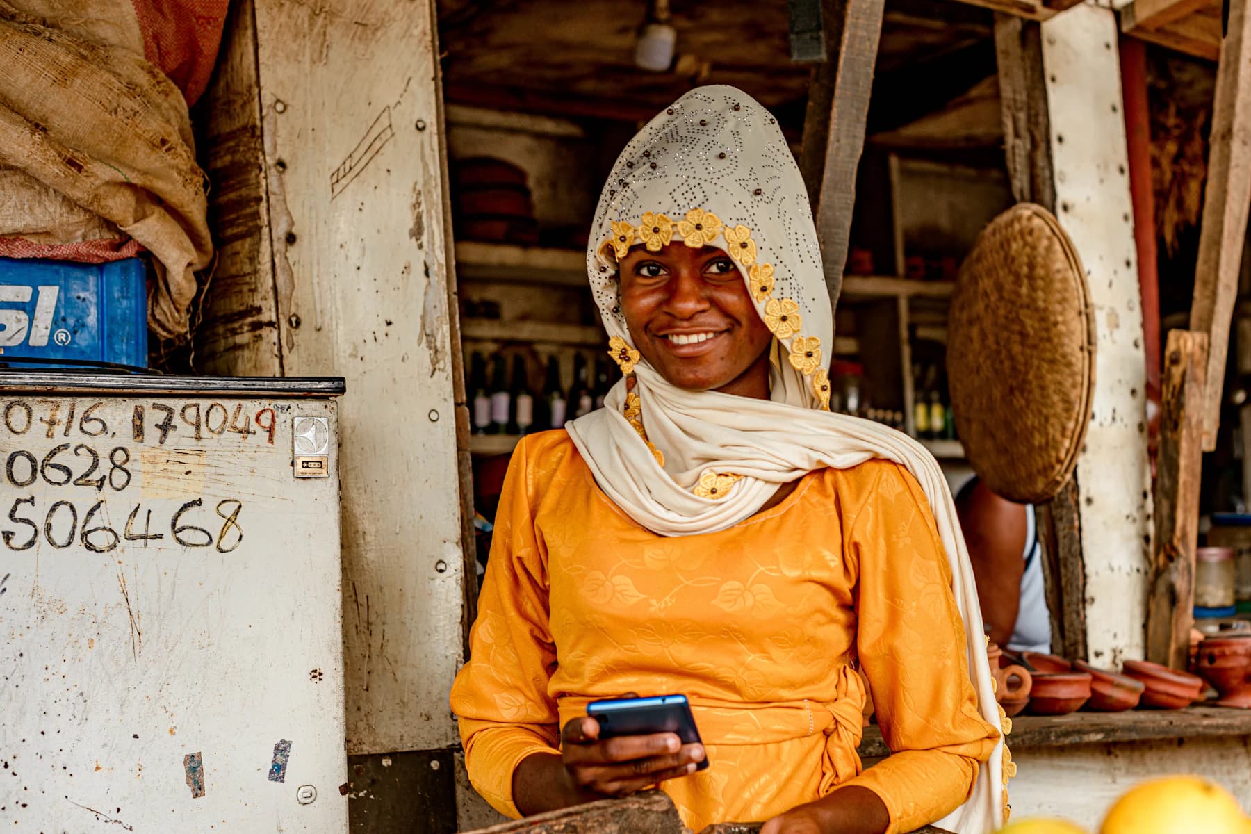 A shop owner in a bright orange dress smiles while checking her phone behind the counter of her roadside shop